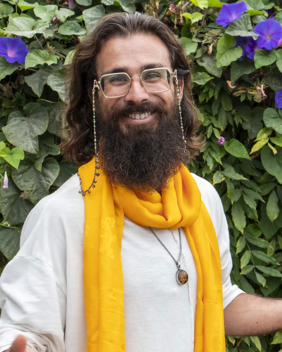 Awe in white with a marigold scarf, smiling with an open hand, morning glories in bloom behind him